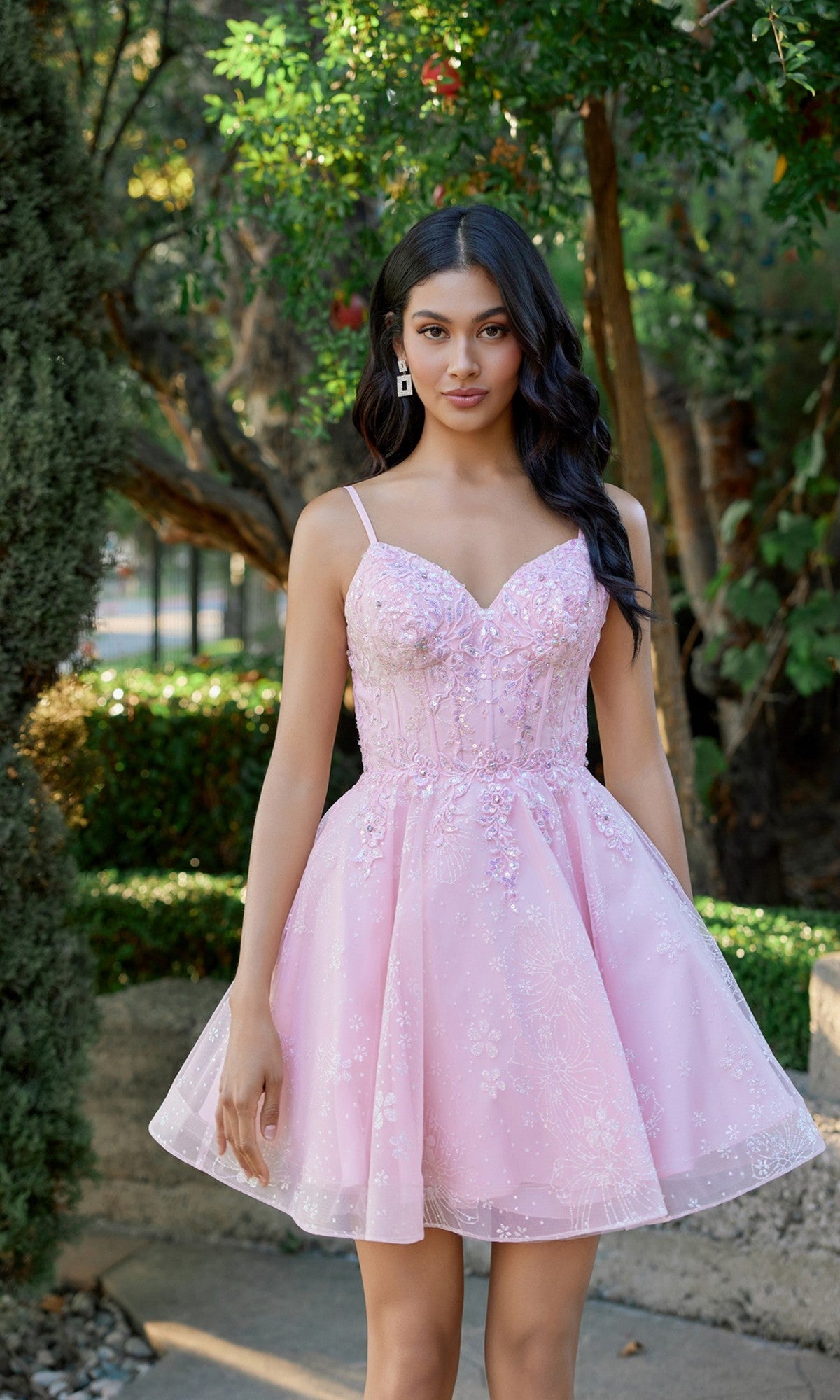 A woman stands outdoors surrounded by greenery and trees, wearing the Juliet JT985L short homecoming dress—a light pink, sleeveless design featuring floral embroidery and a delicate corset bodice.